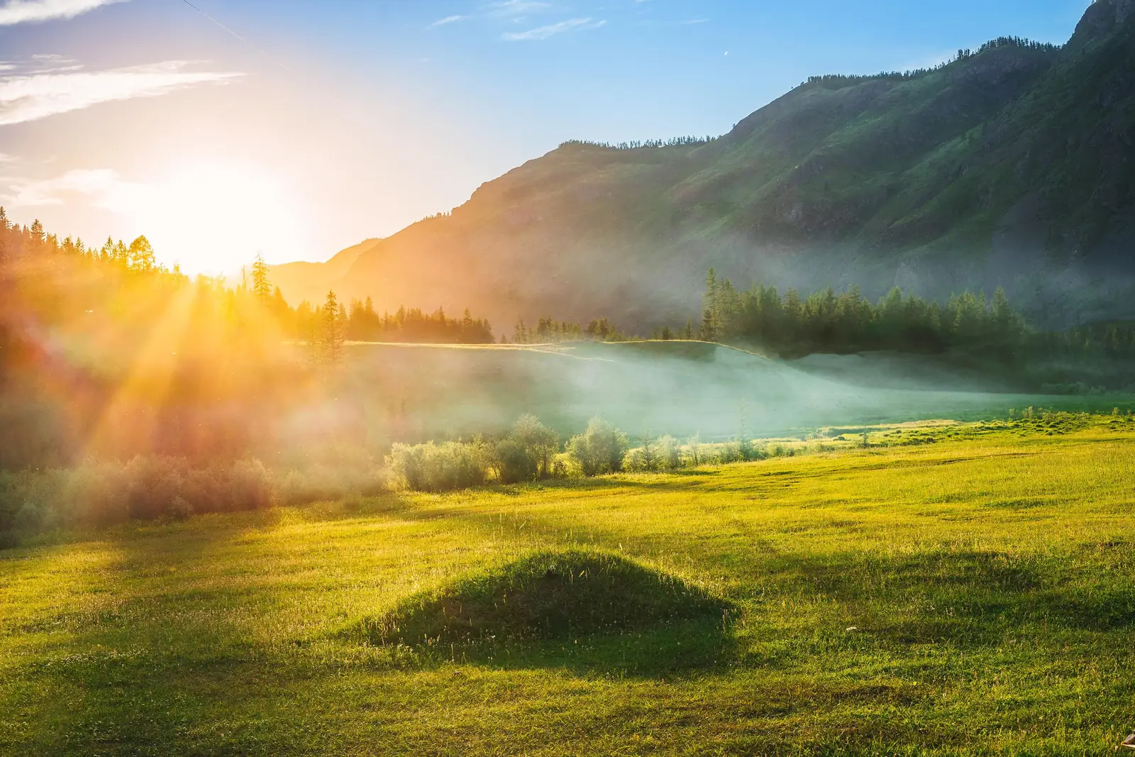 Sun rising over an open green mountain valley as morning mist clears